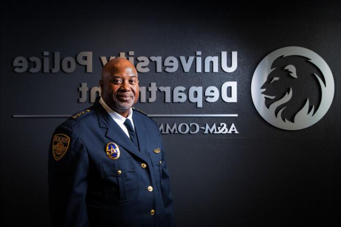 Chief Bryan Vaughn dressed in his dark blue, formal police uniform. He is standing in front of a dark wall with a silver embossed TAMUC lion head logo and the words University Police Department A&移动商务.