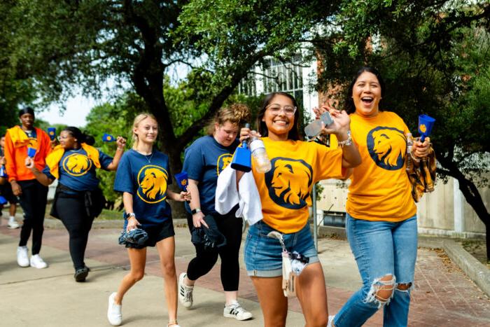 Students walk down the sidewalk on campus, smiling at the camera. Two students in front wear gold t-shirts with the school's lion logo on the front. The students are flashing the "L" hand sign as a show of school spirit.