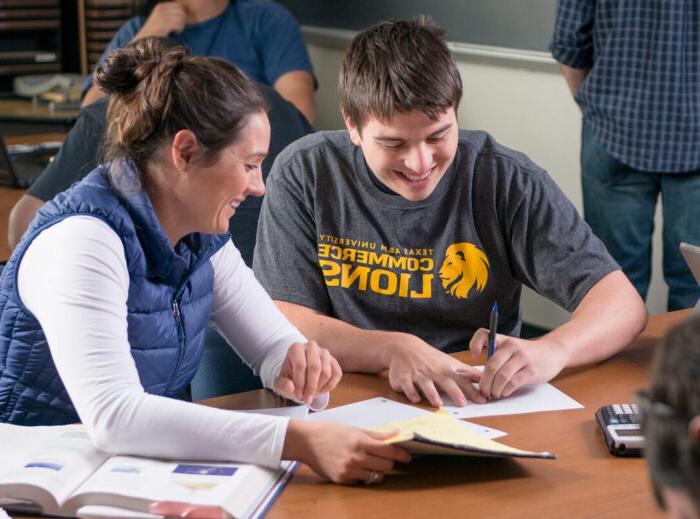 Two students work together at a table in a classroom. 背景中的一个学生在黑板前.